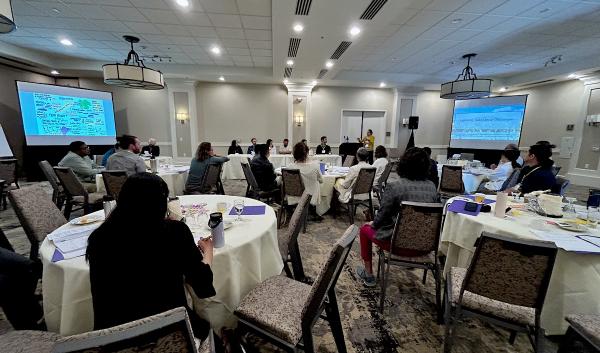 A large conference room with tables of participants facing a group of panelists.