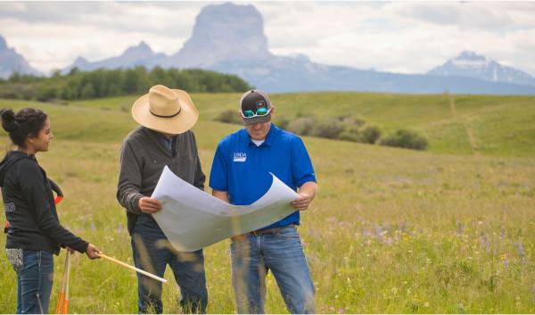 People looking at a map on a rangeland landscape with trees and rugged hills in the background.