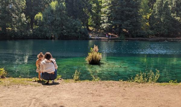 : A mother and young daughter face away from the camera, looking into a blue-green lake. The mother crouches next to her daughter. Across the lake are tall evergreen trees.